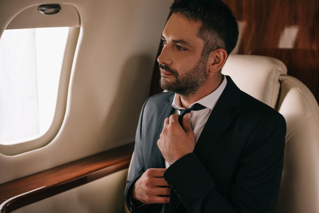 bebaarde zakenman aanraken das en zitten in het vliegtuig - Foto, afbeelding