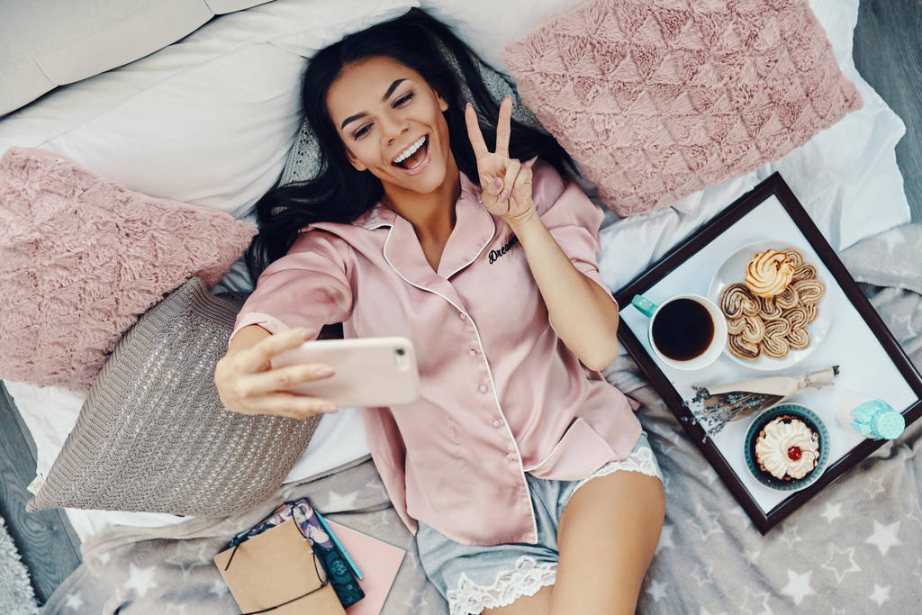 Top näkymä kaunis nuori nainen pyjamassa ottaen selfie ja hymyillen nukkuessaan sängyssä kotona
             - Valokuva, kuva