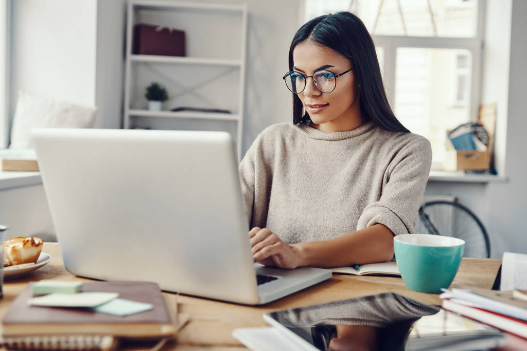 Mulher bonita em roupas casuais usando laptop e sorrindo enquanto trabalhava dentro de casa - Foto, Imagem