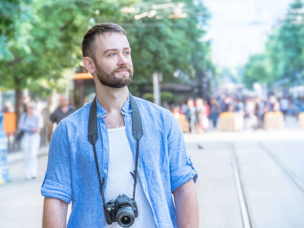 Portrait d'un jeune homme beau caucasien avec une caméra sur fond urbain. La trentaine, barbu, cheveux foncés et look décontracté. Espace de copie
. - Photo, image