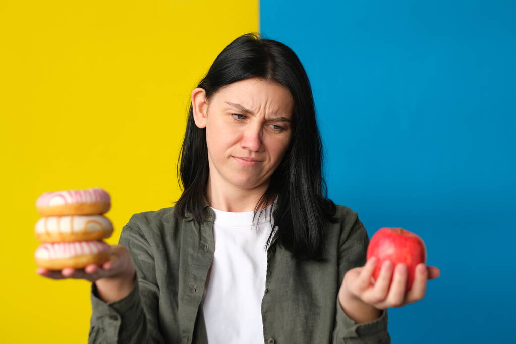 besluiteloze vrouw met donuts en verse rode appel op blauwe en gele achtergrond - Foto, afbeelding