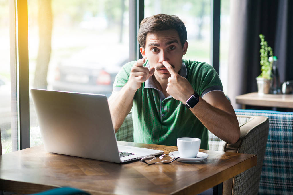 Young angry businessman in green t-shirt pointing at camera and touching his nose while showing lie gesture and sitting at table with laptop in cafe, business and freelancing concept - Fotoğraf, Görsel