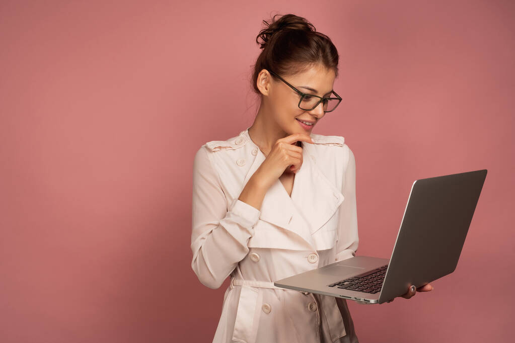 mooi meisje in een licht jasje en bril kijkt in een open laptop in haar handen, staan op een roze achtergrond. - Foto, afbeelding