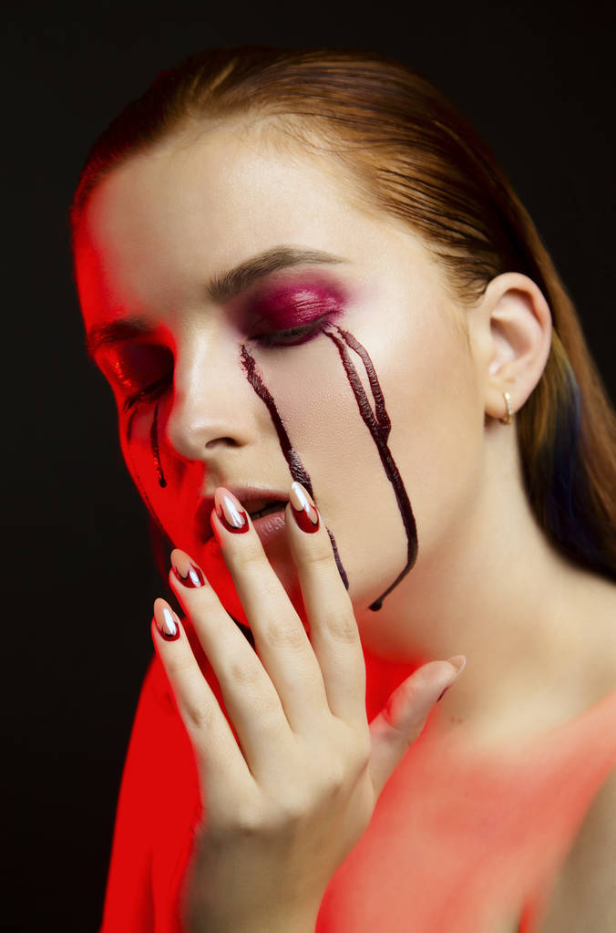 Страшная красота Хэллоуин макияж портрет красивой рыжей девушки с красными тенями для глаз и фальшивой кровью от желтых глаз, студийный портрет, красный свет, черный фон
 - Фото, изображение