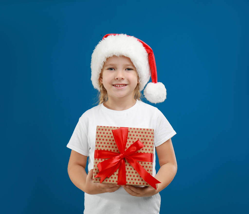 niedliches kleines Kind trägt Weihnachtsmütze mit Weihnachtsgeschenk auf blauem Hintergrund - Foto, Bild