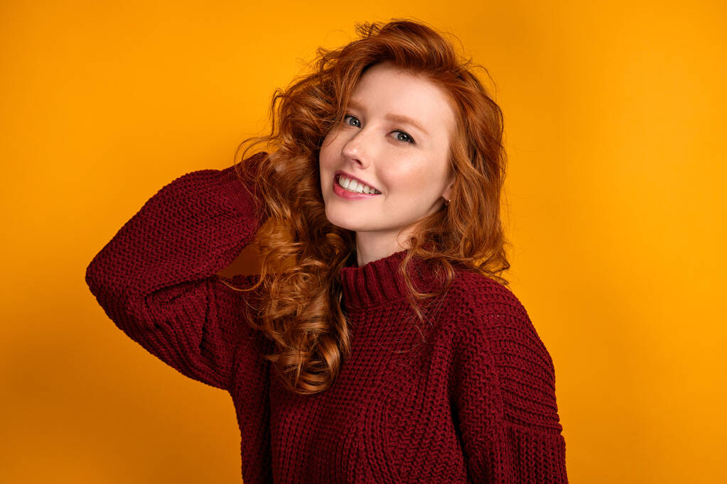 Redhead curly girl in a burgundy sweater stands straightens her hair and smiles broadly against a yellow background - 写真・画像
