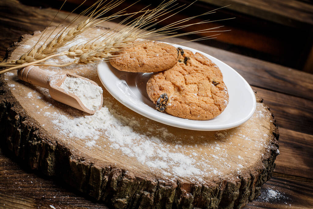 Biscoitos de gengibre de aveia com passas, farinha e espigas de trigo em uma superfície de madeira
 - Foto, Imagem