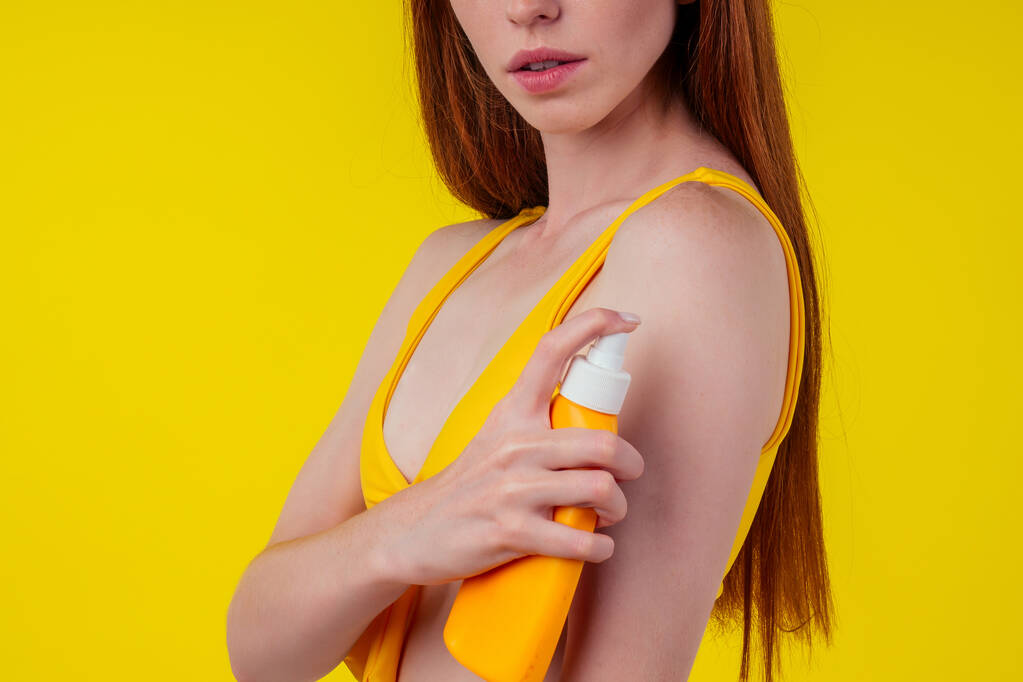beautiful edheaded ginger woman applying spf sun rays protetor on shoulders arms and neck wearing stylish bikini in studio yellow background - Photo, Image