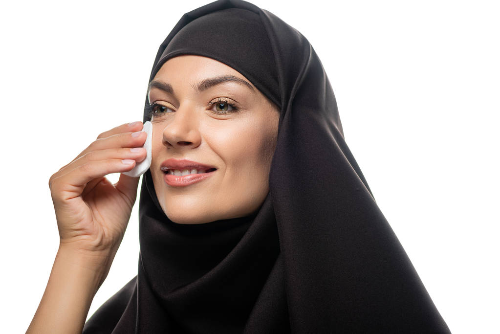 χαμογελαστή νεαρή μουσουλμάνα γυναίκα σε μαντίλα σκούπισμα πρόσωπο με βαμβάκι μαξιλάρι απομονώνονται σε λευκό - Φωτογραφία, εικόνα