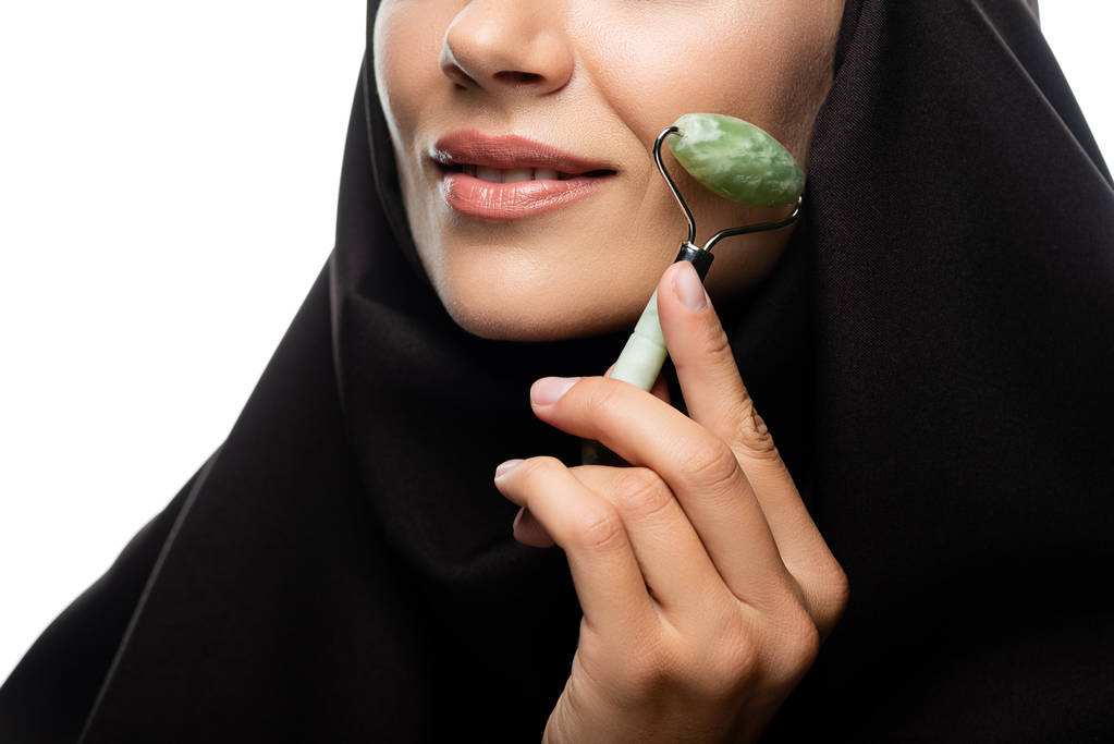 vista parcial de la joven musulmana en hijab usando rodillo de jade aislado en blanco
 - Foto, Imagen