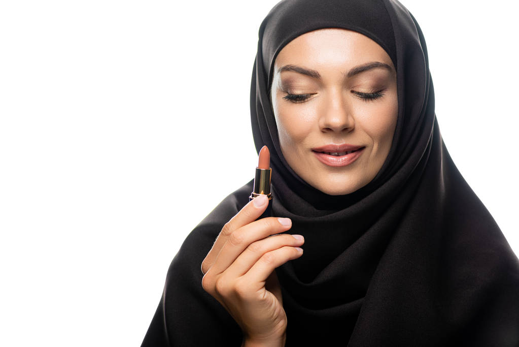 χαμογελαστή νεαρή μουσουλμάνα γυναίκα με μαντίλα που κρατά το μπεζ κραγιόν απομονωμένο σε λευκό - Φωτογραφία, εικόνα