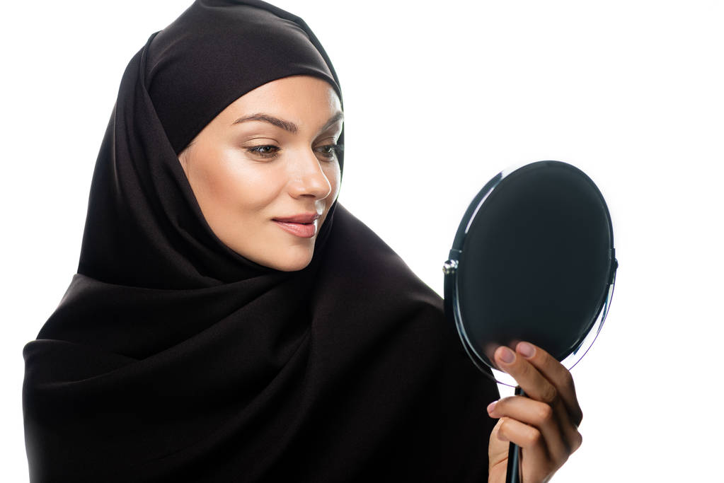 νεαρή μουσουλμάνα γυναίκα με μαντίλα κοιτάζει τον καθρέφτη που είναι απομονωμένος στα λευκά - Φωτογραφία, εικόνα