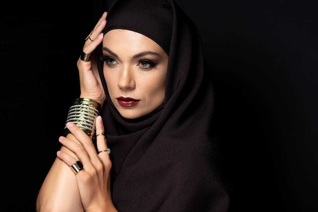 красивая мусульманка в хиджабе с макияжем в золотых кольцах и браслетом изолированы на черном
 - Фото, изображение