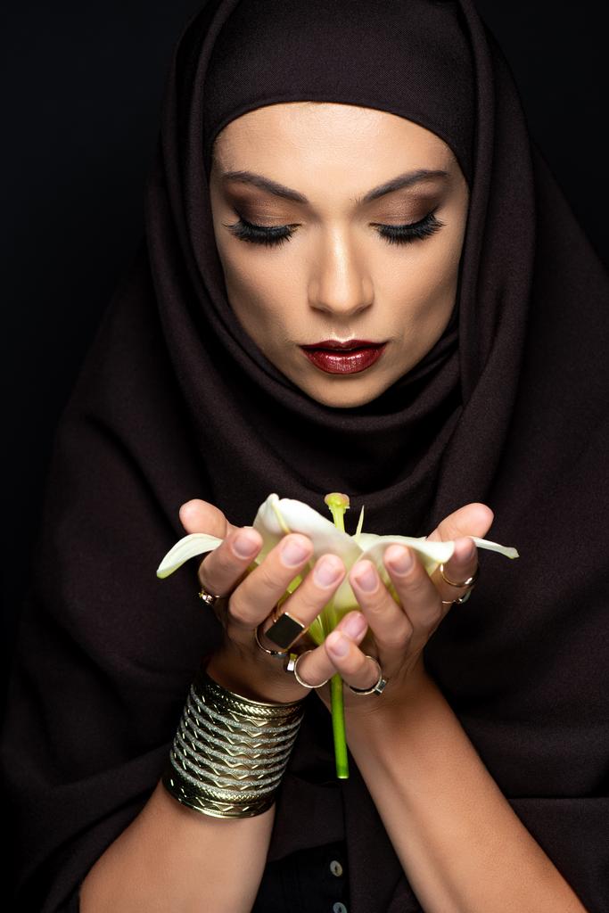 beautiful Muslim woman in hijab in golden rings and bracelet holding lily isolated on black - Photo, Image