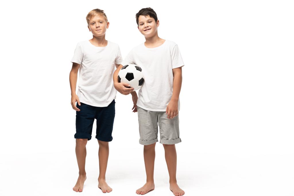 Neşeli çocuk futbol topunu tutarken gülümseyen kardeşinin yanında beyaz arka planda - Fotoğraf, Görsel