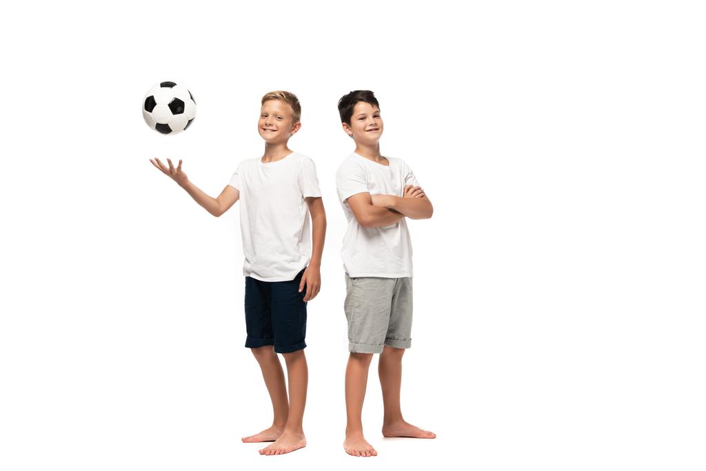 Gülümseyen kardeşin yanında futbol oynayan neşeli çocuk beyaz arka planda çapraz kollarla duruyor. - Fotoğraf, Görsel