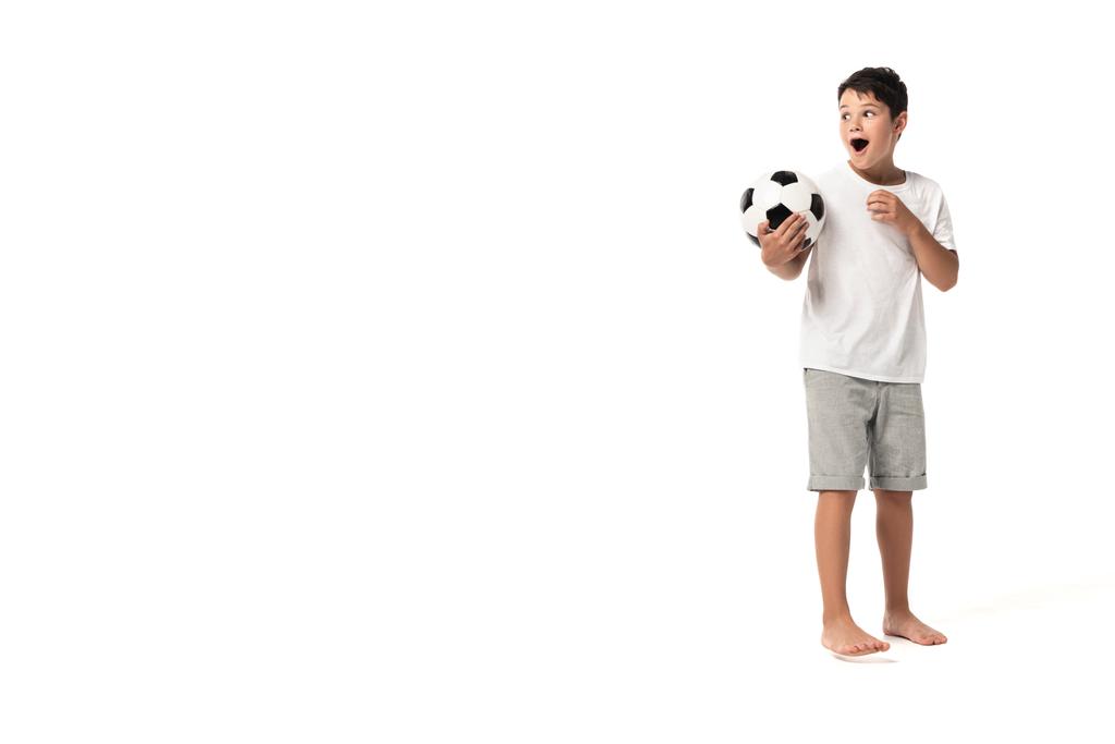 překvapený chlapec drží fotbalový míč, dívá se jinam a ukazuje prstem na bílém pozadí - Fotografie, Obrázek