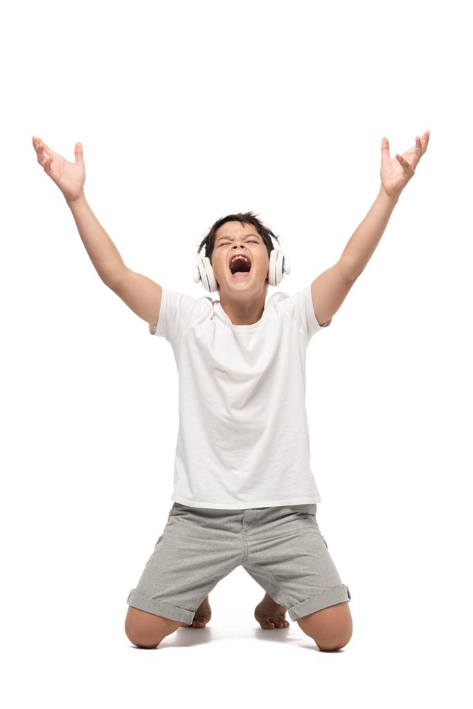 excited boy standing on knees with raised hands while listening music in headphones and singing on white background - Photo, Image