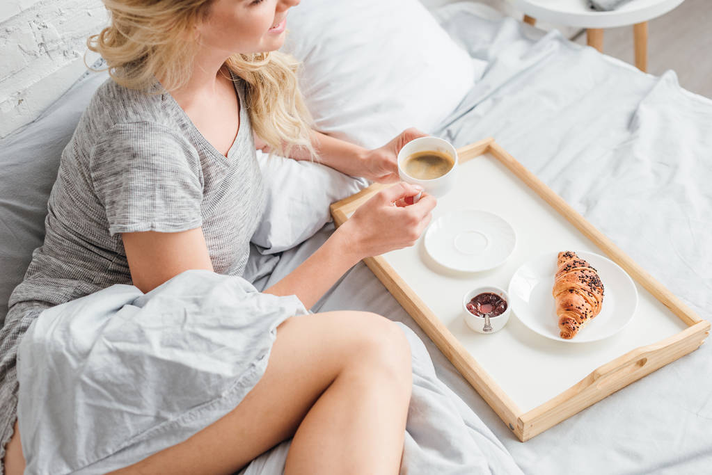 vista recortada de mujer alegre sosteniendo taza de café cerca de bandeja de madera con croissant en la cama
  - Foto, imagen