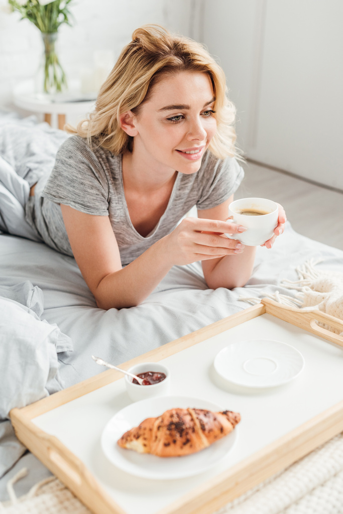 selective focus of cheerful girl holding cup of coffee near tray with tasty croissant on bed  - Photo, Image