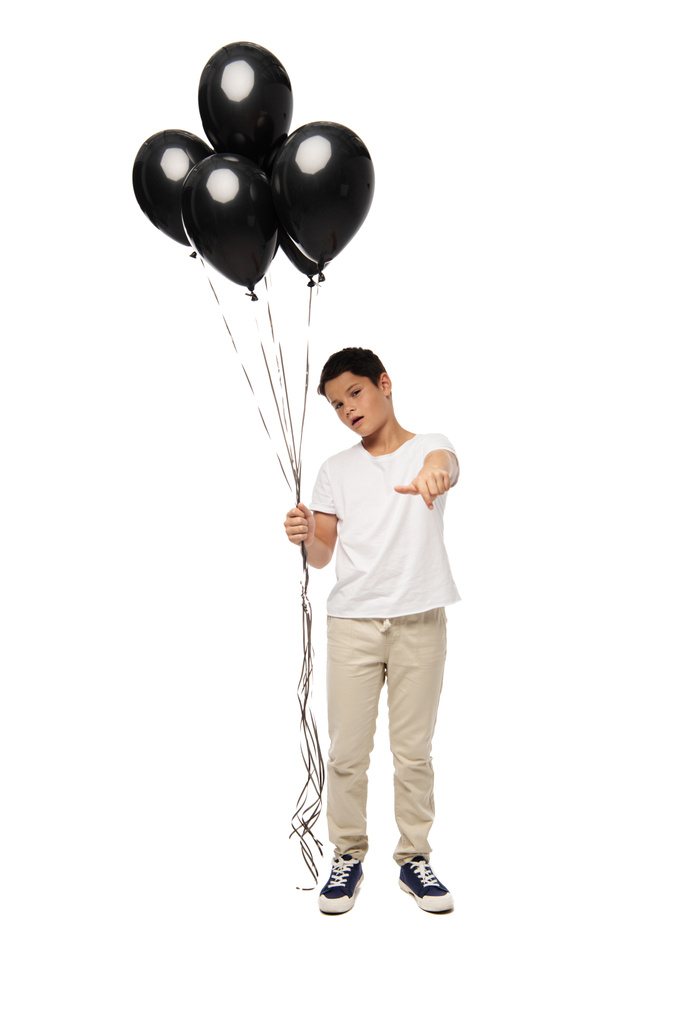 ernster Junge, der mit der Hand auf die Kamera zeigt, während er schwarze Luftballons auf weißem Hintergrund hält - Foto, Bild