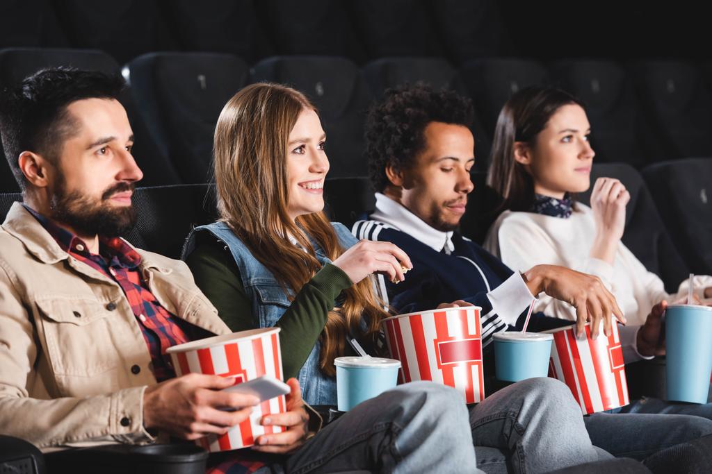 wielokulturowi i uśmiechnięci przyjaciele trzymający popcorn i oglądający film w kinie  - Zdjęcie, obraz