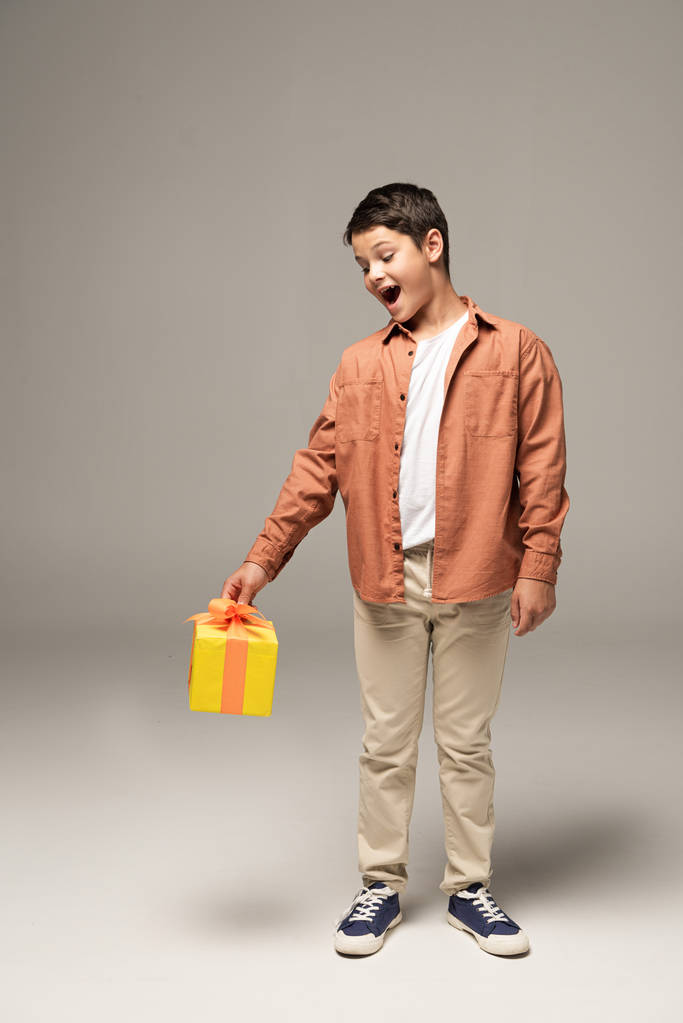 niño emocionado sosteniendo caja de regalo amarillo mientras está de pie sobre fondo gris
 - Foto, Imagen