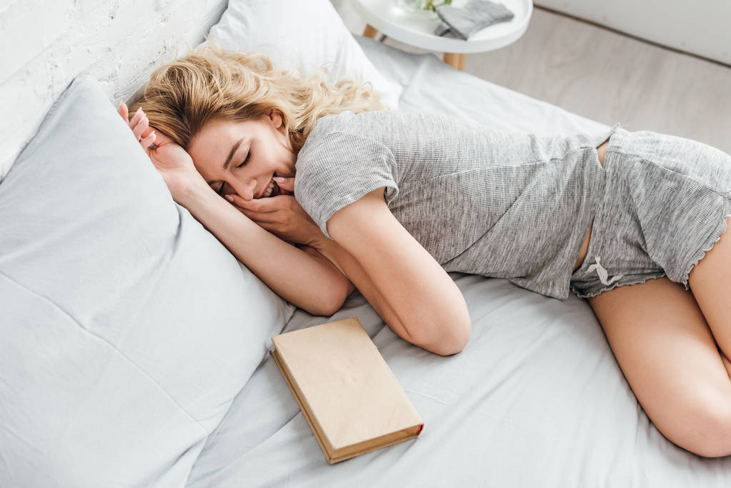 над головой вид веселой женщины, покрывающей лицо смехом и лежащей на кровати возле книги
  - Фото, изображение
