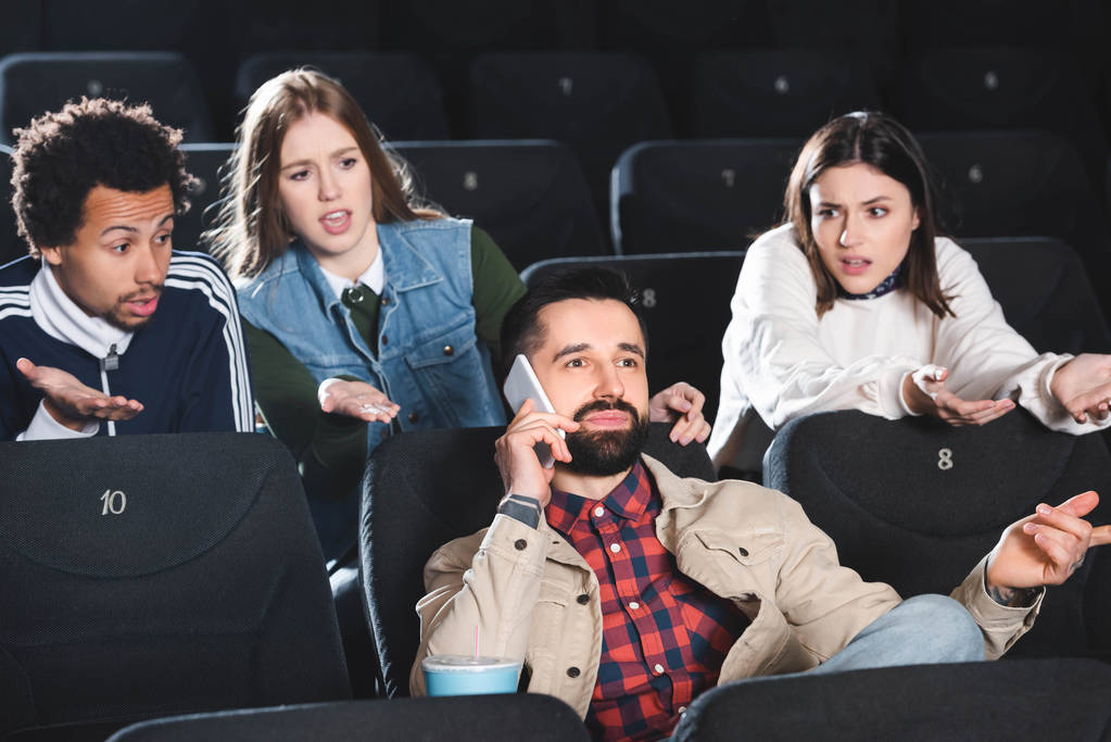 Irritierte multikulturelle Freunde schauen Mann im Kino beim Smartphone-Gespräch an  - Foto, Bild