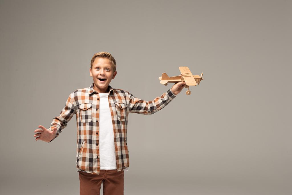 возбужденный мальчик держа деревянный игрушечный самолет и глядя на камеру изолированы на серый
 - Фото, изображение