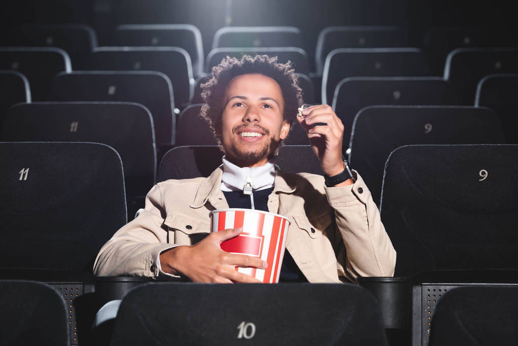 sorridente uomo africano americano che tiene popcorn e guarda film nel cinema
  - Foto, immagini