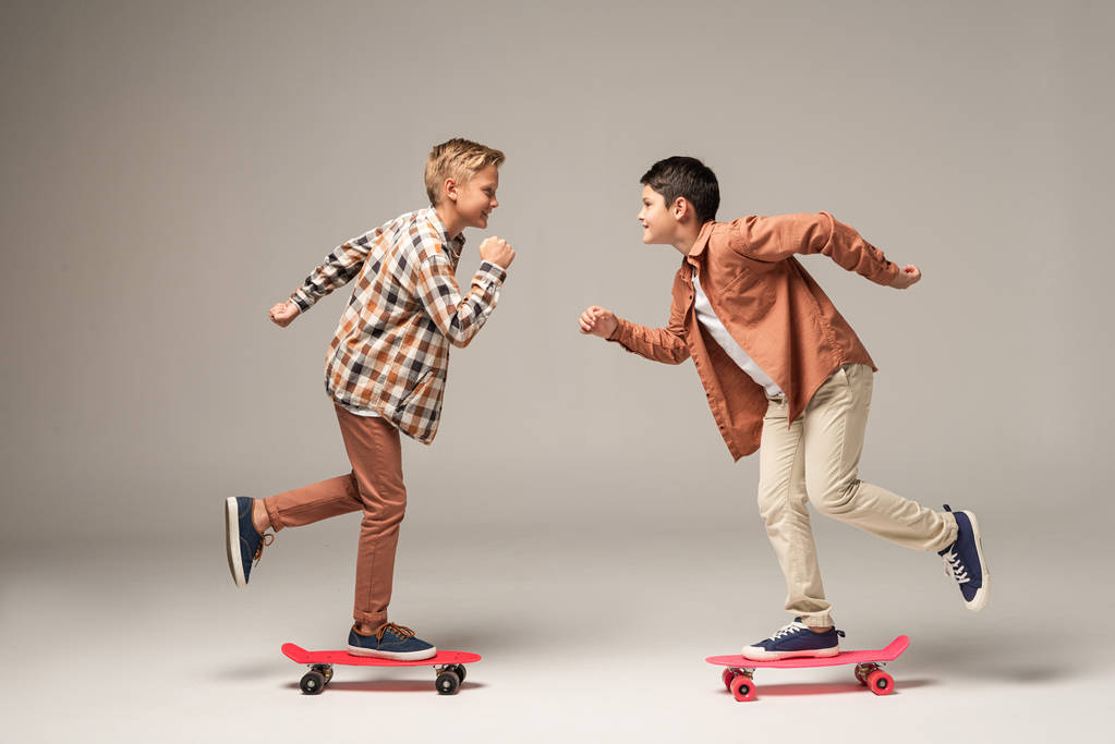 Seitenansicht von zwei fröhlichen Brüdern, die Penny-Boards reiten, während sie einander auf grauem Hintergrund anschauen - Foto, Bild