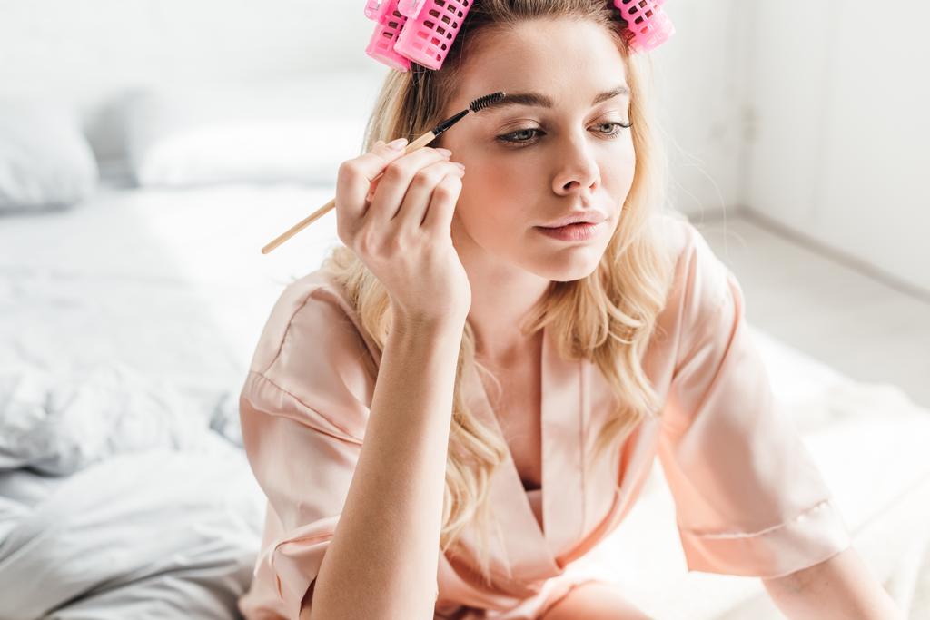 beautiful woman in pink hair curlers styling eyebrows in bedroom  - Photo, Image