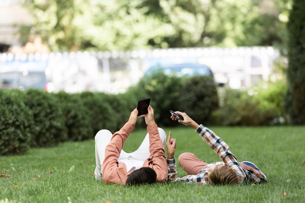 twee broers liggend op groen gazon op de rug en met behulp van smartphones - Foto, afbeelding