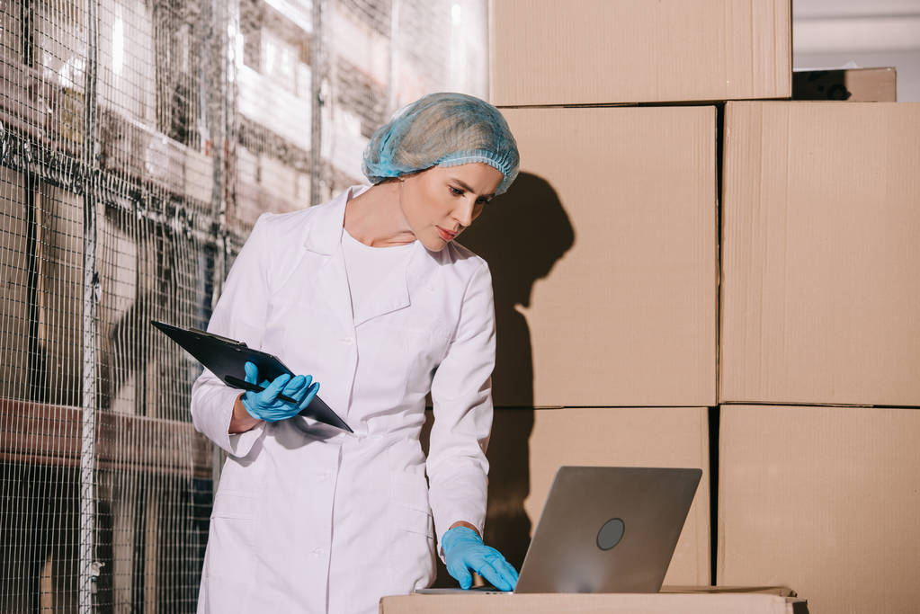 concentrated storekeeper in white coat and hairnet using laptop and holding clipboard  - Photo, Image