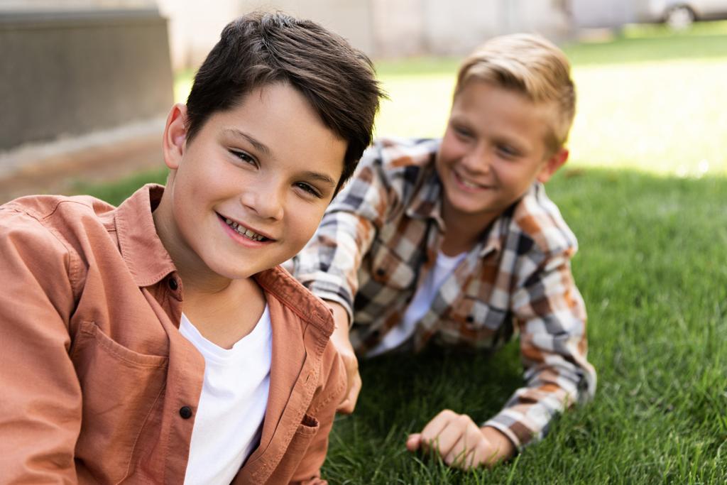 Selektiver Fokus des fröhlichen Jungen, der in die Kamera blickt, während er in der Nähe seines Bruders auf dem Gras liegt - Foto, Bild