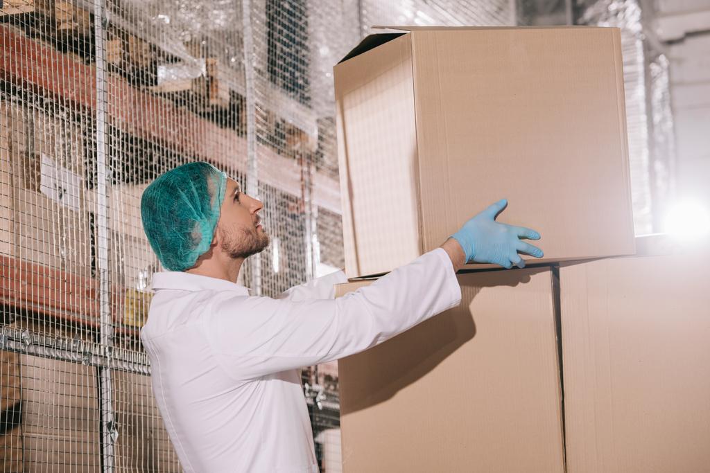 storekeeper in white coat and hairnet taking cardboard box in warehouse - Photo, Image