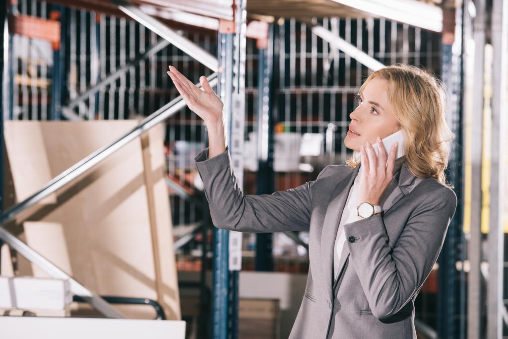serious businesswoman talking on smartphone, looking away and pointing with hand in warehouse - Photo, Image