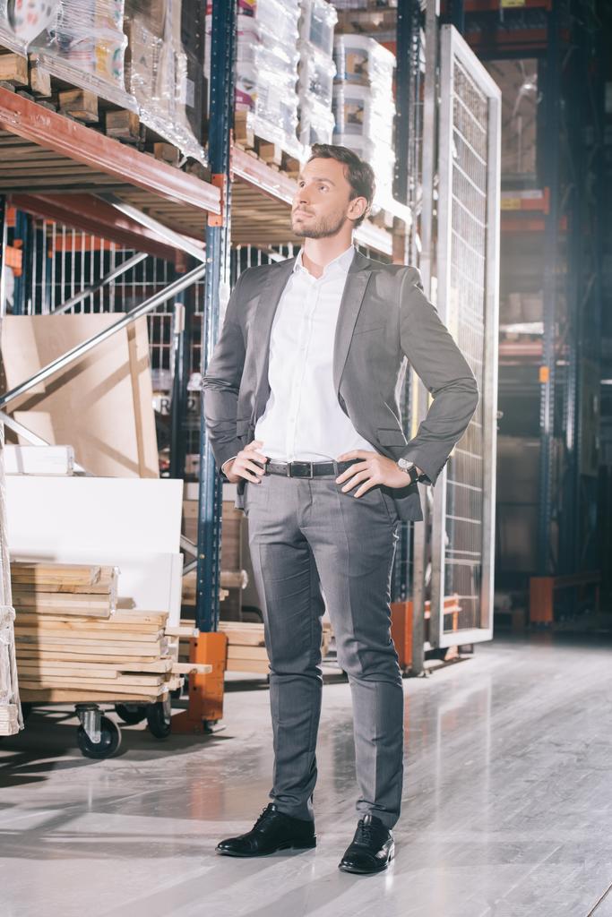 confident businessman looking away while standing in warehouse with hands on hips - Photo, Image