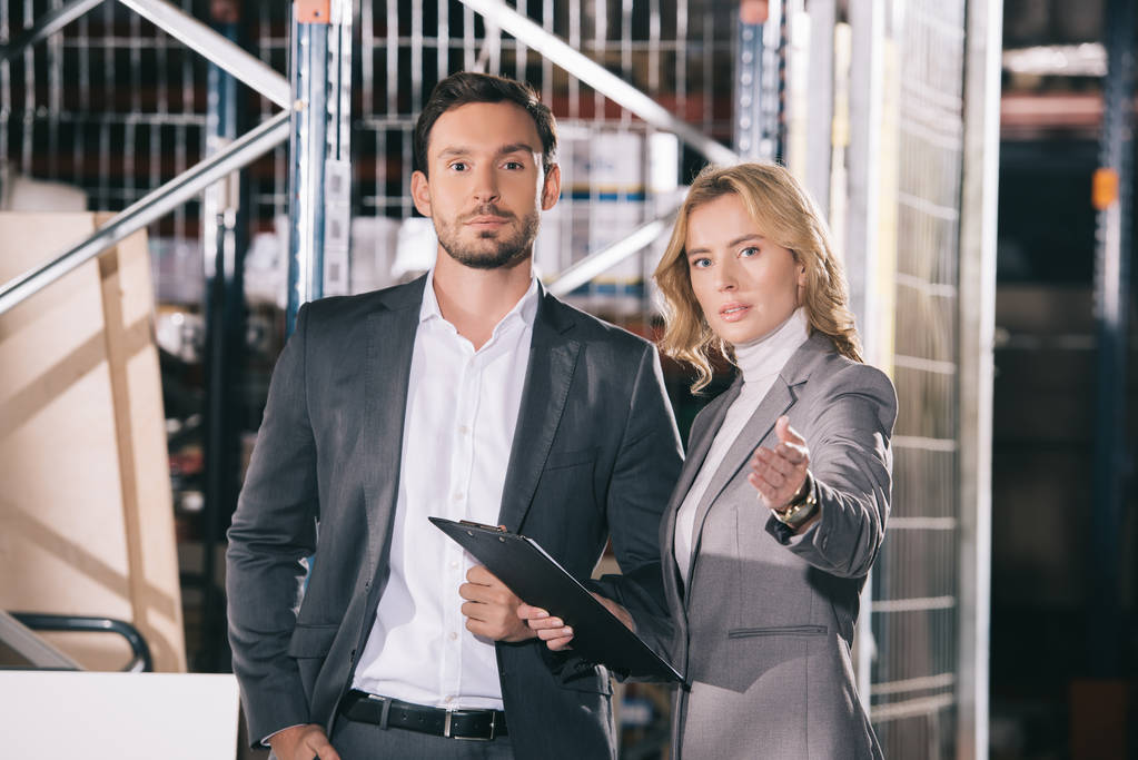attractive businesswoman pointing with hand at camera while standing near confident businessman in warehouse - Photo, Image