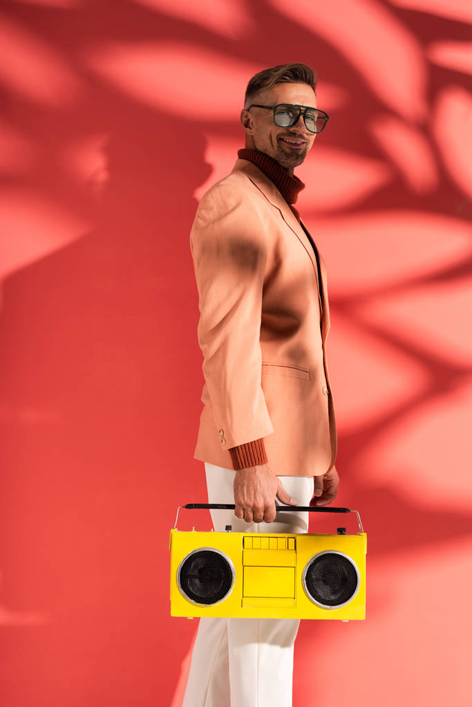 szczęśliwy i stylowy mężczyzna w okularach przeciwsłonecznych trzyma żółty boombox na czerwono z cieniami  - Zdjęcie, obraz