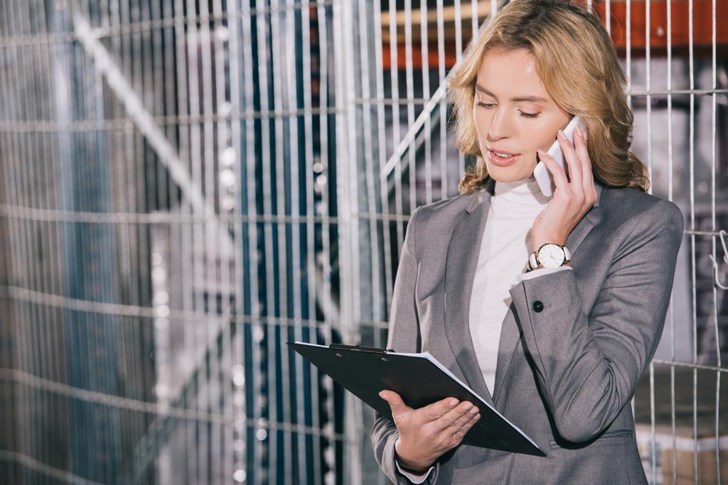 atractiva mujer de negocios hablando en el teléfono inteligente y sujetando portapapeles en el almacén
 - Foto, imagen