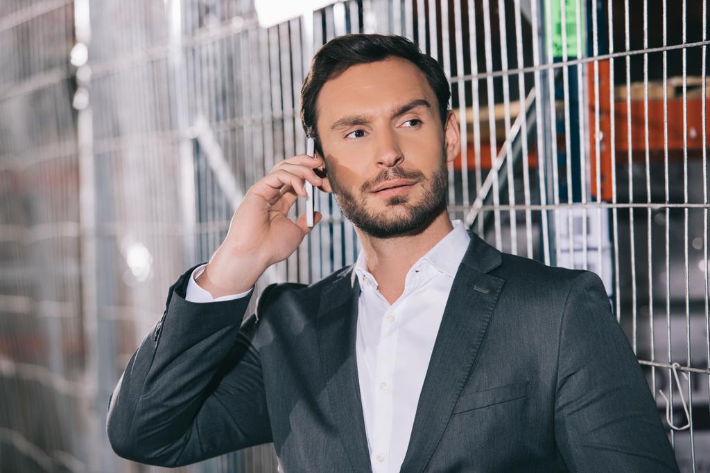 confident businessman looking away while talking on smartphone in warehouse - 写真・画像
