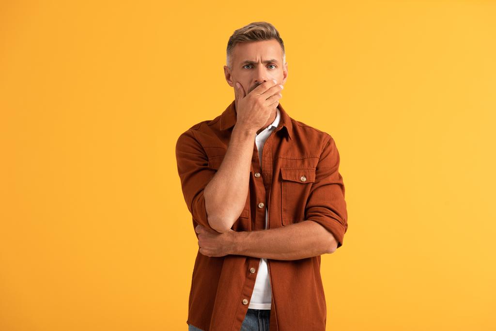 rozrušený muž zakrývá ústa a dívá se na kameru izolované na oranžové  - Fotografie, Obrázek