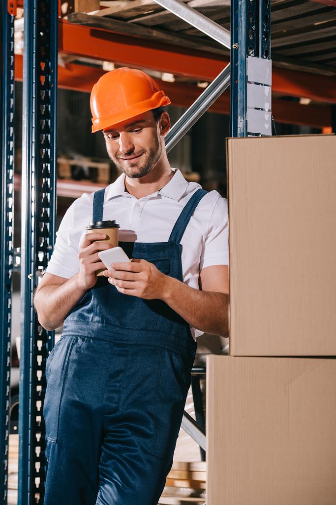 smiling loader holding coffee to go and using smartphone in warehouse - 写真・画像