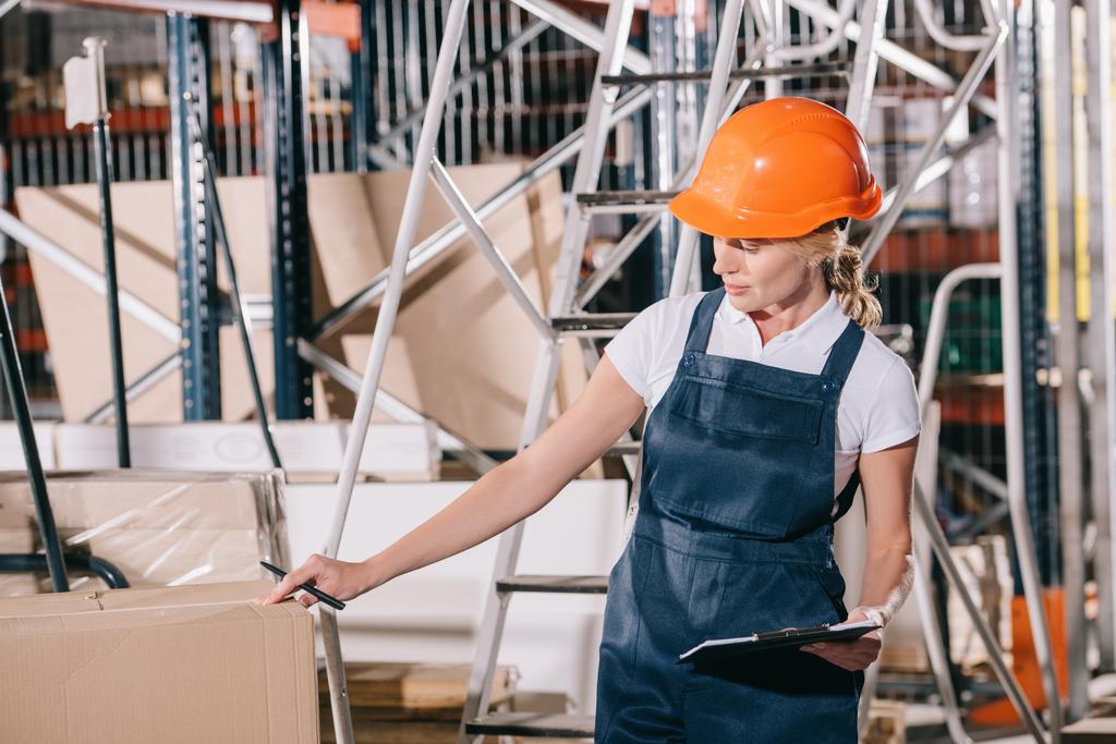 workwoman v montérkách a helmě držící schránku při kontrole kartonové krabice - Fotografie, Obrázek