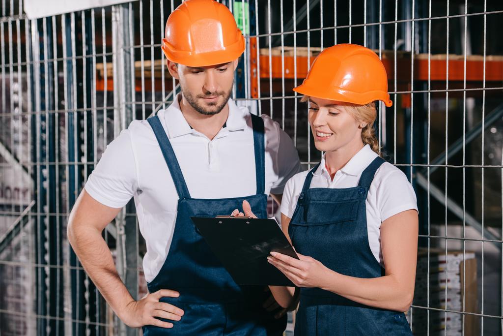 sonriente trabajadora sosteniendo portapapeles mientras está de pie cerca de trabajador concentrado
  - Foto, Imagen