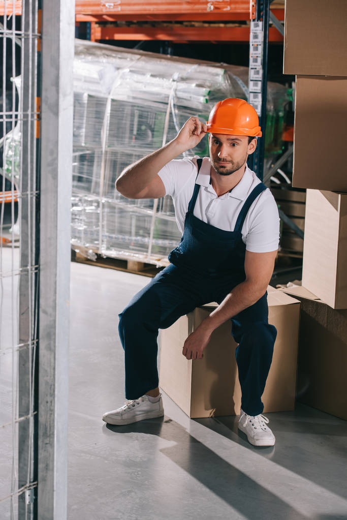 tired loader sitting on cardboard box in wearhouse and touching helmet - Photo, Image