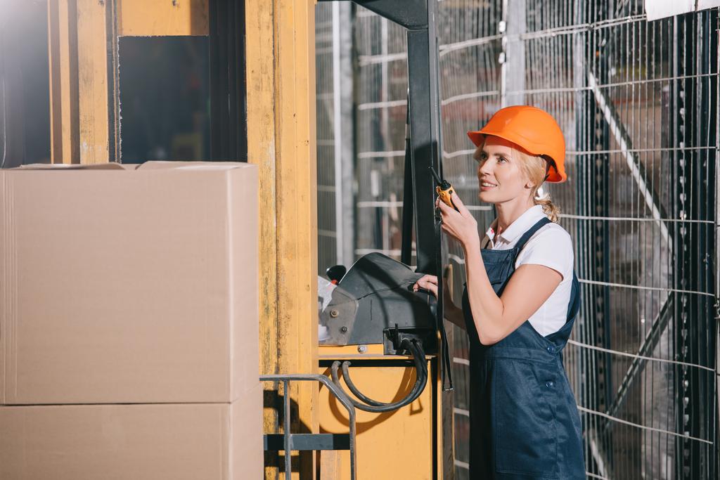 улыбающаяся работница разговаривает по рации, стоя рядом с погрузчиком
 - Фото, изображение