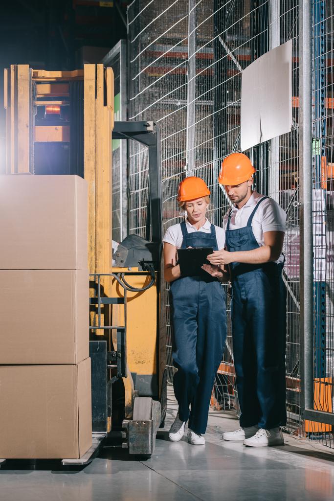 フォークリフトローダーの近くに立っている間にクリップボードを見ている2人の倉庫労働者 - 写真・画像